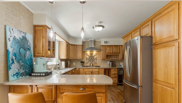 A kitchen with wooden cabinets, a stainless steel refrigerator, a marble countertop, and two bar stools. - Tulsa Turnaround 4 bed (Tulsa)