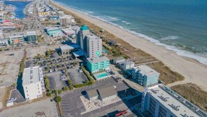 An aerial view of a coastal town with a prominent hotel featuring a blue roof, adjacent to a beach and ocean. - Tidewinds 202 - Magnolia Sand at The Boardwalk (Carolina Beach)