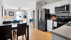 A modern kitchen with stainless steel appliances, a dining area with dark chairs, and a view of the outdoors. - Tidewinds 202 - Magnolia Sand at The Boardwalk (Carolina Beach)