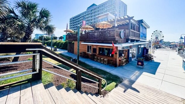 A beachfront hotel with a wooden deck, outdoor seating, and a view of the ocean. - Tidewinds 202 - Magnolia Sand at The Boardwalk (Carolina Beach)
