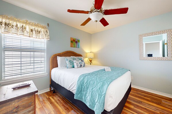Une chambre à coucher comprenant un lit, un ventilateur de plafond, une fenêtre avec des stores, une table de chevet et une commode.