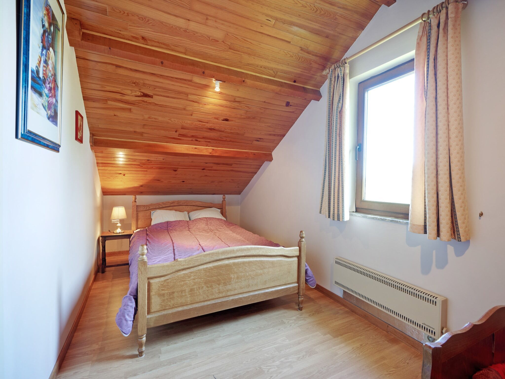 A bedroom with a wooden ceiling, a bed, a nightstand, a window with curtains, and a radiator.