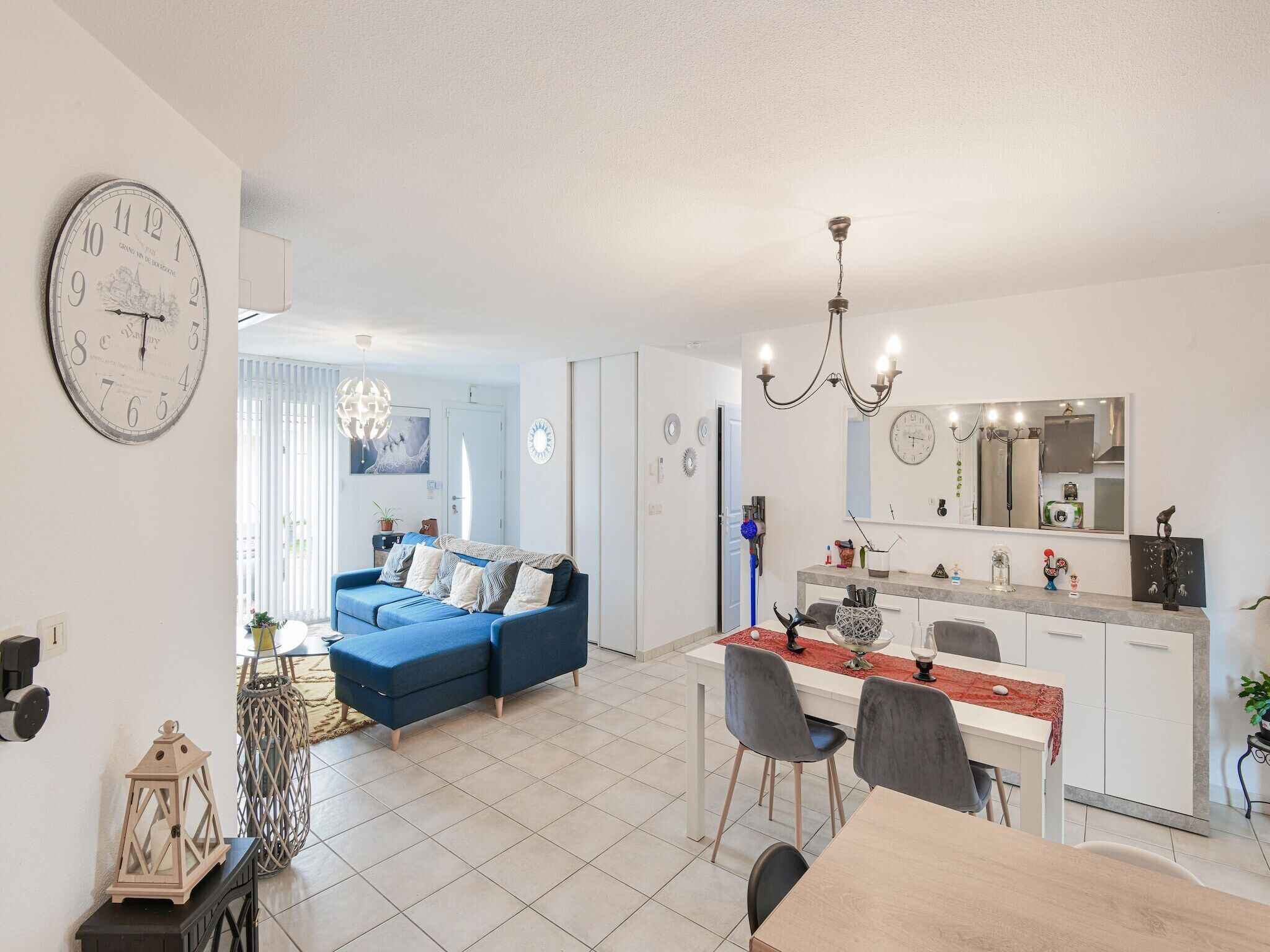 A modern living room with a blue sofa, a dining area with a white table, and a kitchen with a large mirror. - Zen room in shared home with pool (owner-on site) (Saint-Médard de Guizières)