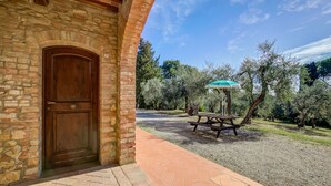 Un edificio rústico de piedra con puerta de madera, un camino empedrado y una mesa de picnic con sombrilla en un parque con olivos.