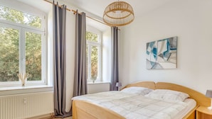 A bedroom with a wooden bed, large windows with curtains, a rattan light fixture, and a painting on the wall.