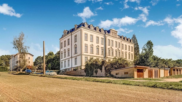 A large, multi-story building with a symmetrical facade, surrounded by a field and trees.
