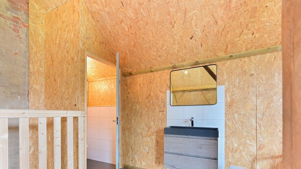 A room with a wooden floor, a white wall with a mirror, a sink, and a trash bin.