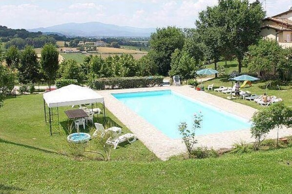 Piscina com toldo branco, cercada por grama e árvores, com vista para colinas ao longe.