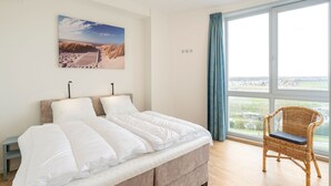 A bedroom with a bed, a wicker chair, a window with a view, and a painting on the wall. - Beautiful apartment with sea view (Scherpenisse)