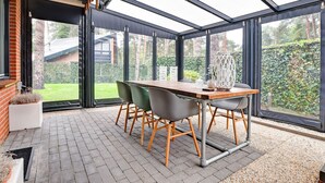 A conservatory with a wooden table and chairs, large windows, and a view of a garden. - Charming holiday home with a covered terrace (Meijel)