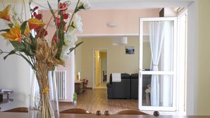 Een eetgedeelte met een tafel, een vaas met bloemen, een radiator en uitzicht op een woonkamer door een geopende deur.