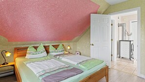 Una camera da letto con soffitto a falde, un letto con biancheria dai motivi decorativi, una lampada da comodino e una porta che dà su un'altra stanza.