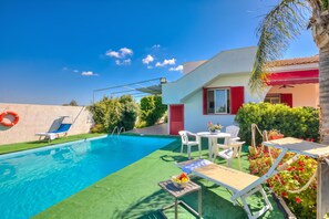 A swimming pool with a sunshade, lounge chairs, and a table with a fruit bowl.