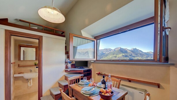 A dining area with a table set for two, a bowl of fruit, and a view of mountains through a large window. - Studio Placide Mountain View, Sauze d'Oulx, Italy (Sauze d'Oulx)