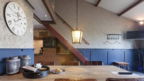 Un coin repas avec une table en bois, des chaises et une grande horloge murale. Un escalier mène à un autre niveau, et un luminaire suspendu éclaire l’espace.