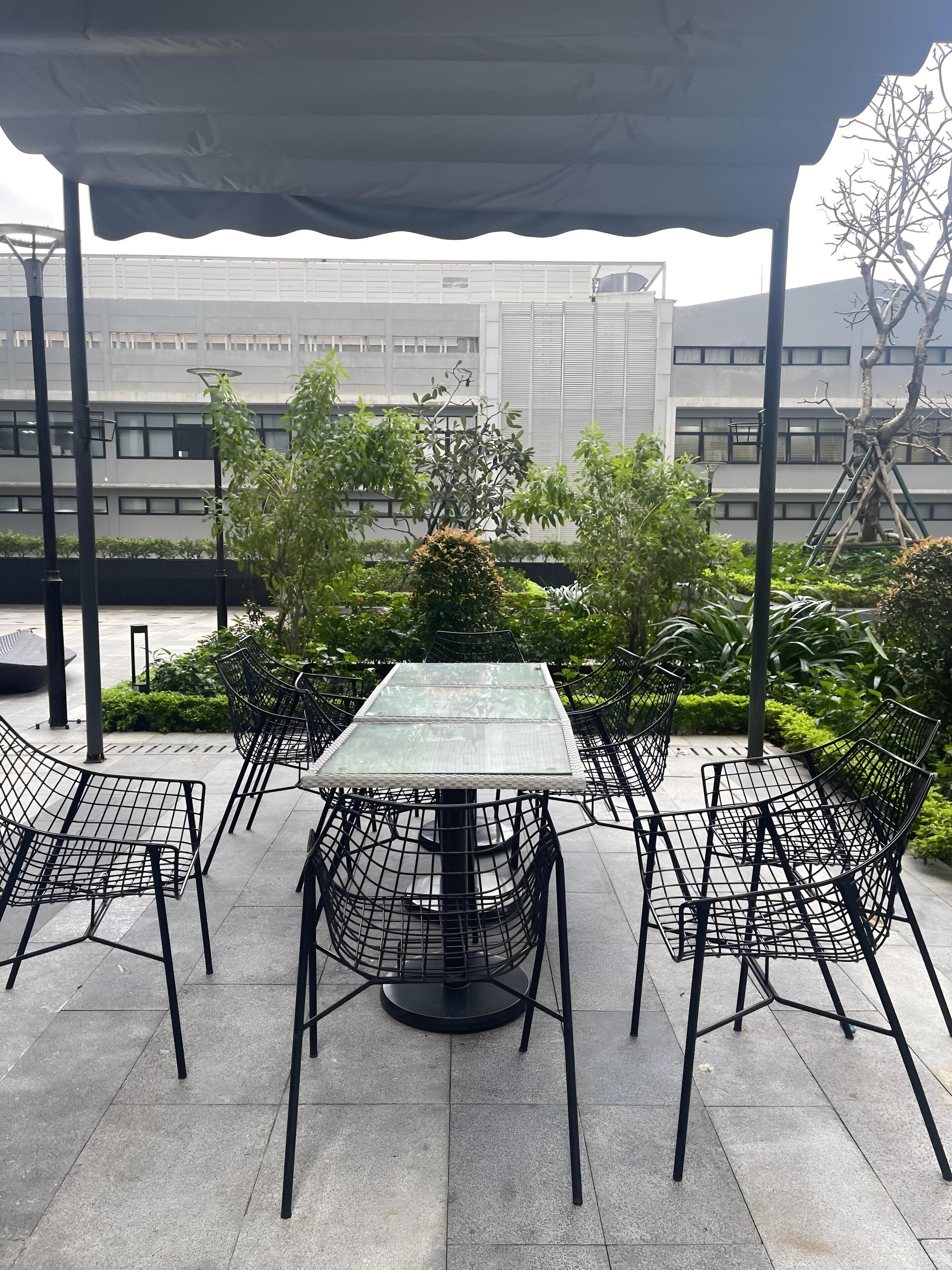 An outdoor patio with a glass-top table and metal chairs, surrounded by greenery and a building in the background.