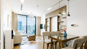 A modern hotel room with a dining area, a sofa, a coffee table, and a large window offering a city view. - Vinhomes Metropolis Ha Noi-Gem Apartment (Hanoi)