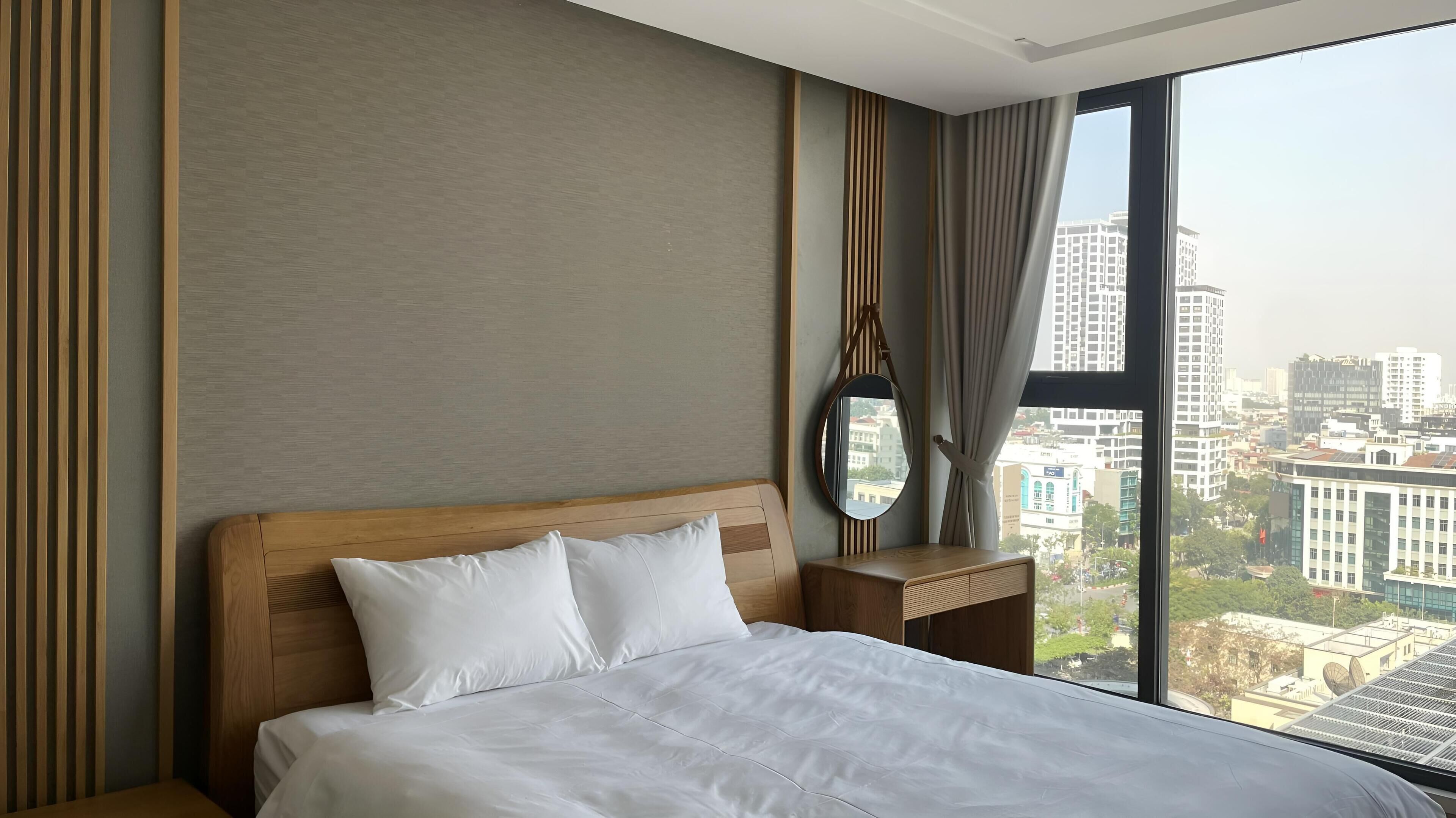 A hotel room with a large bed, a wooden headboard, a bedside table with a mirror, and a view of the city through a large window.