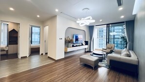 A modern living room with a TV, sofa, and ottoman. There is a wooden floor and a large window with curtains. - Vinhomes Metropolis Ha Noi-Gem Apartment (Hanoi)