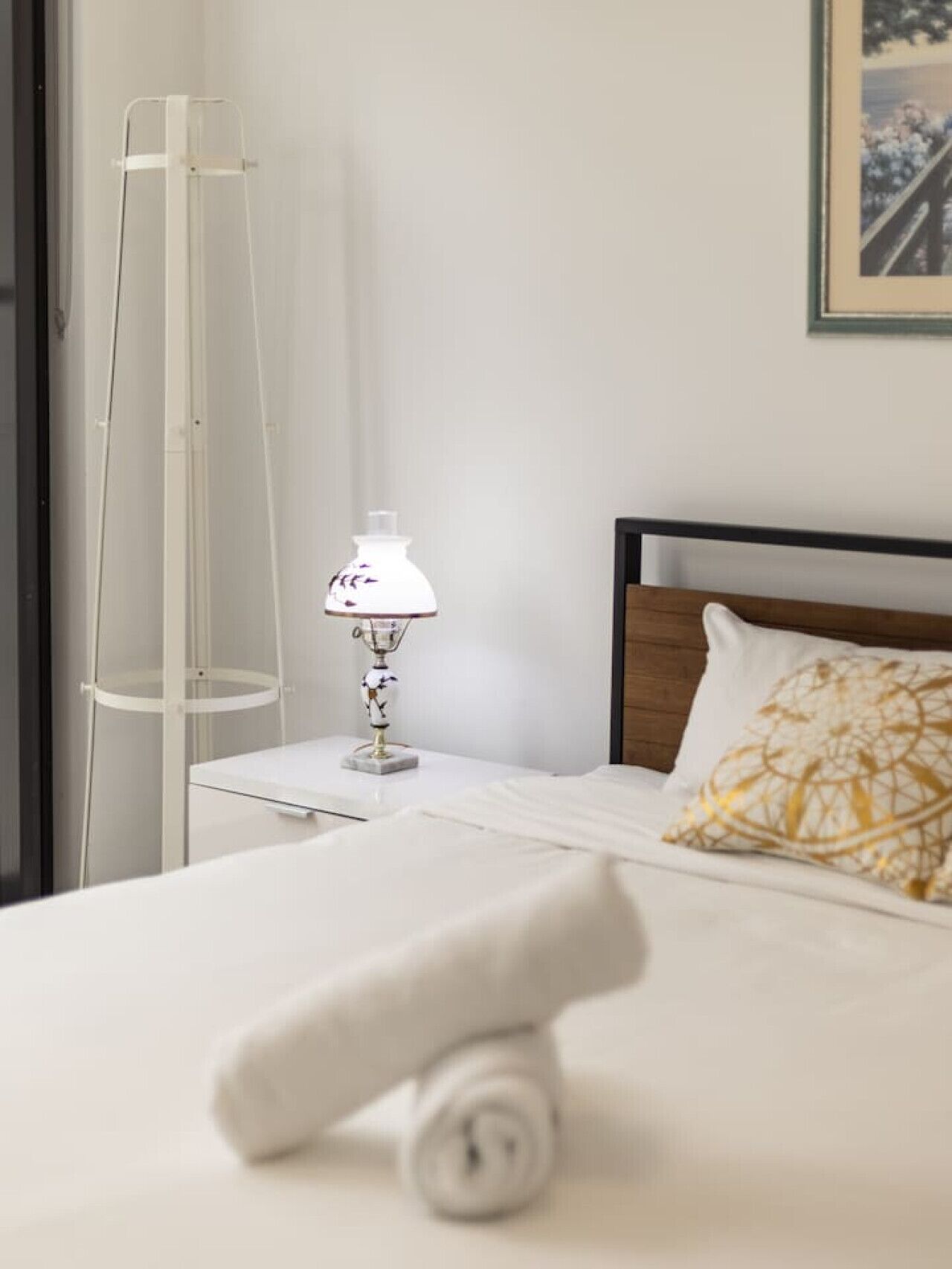 Un lit bien fait, avec un oreiller blanc et une serviette enroulée, une table de chevet avec une lampe et une étagère haute et transparente.