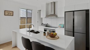 A modern kitchen with white cabinetry, a stainless steel refrigerator, and a central island with a sink and a bowl of fruit. - Brand-new 4BR modern family home @Park Ridge (Park Ridge)