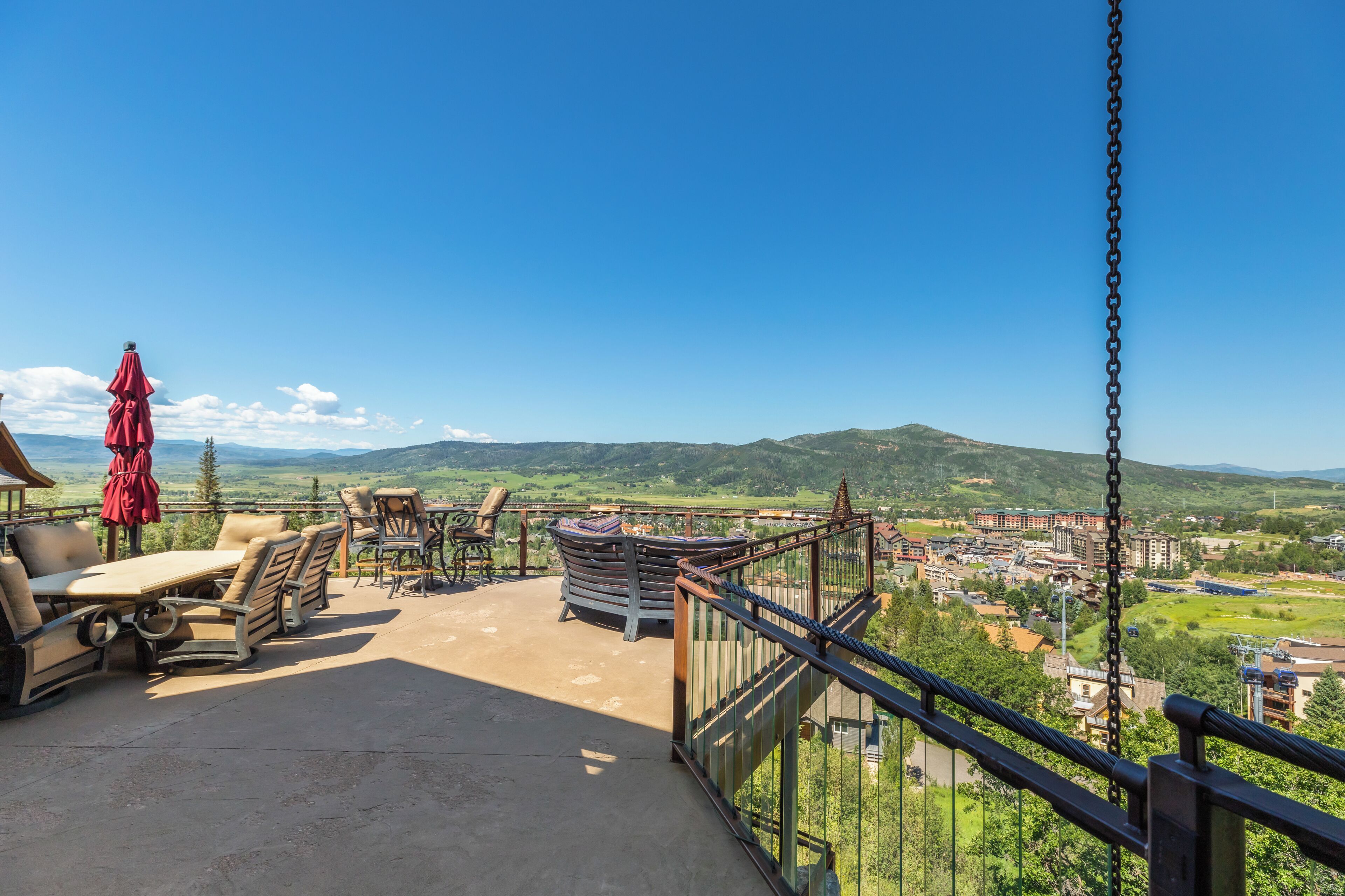 A balcony with outdoor furniture, a red umbrella, and a view of a town and hills.