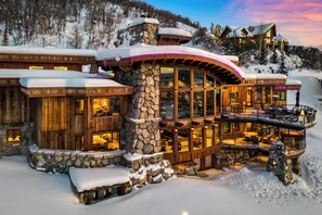 A large, multi-story lodge with stone and wood construction, featuring large windows and a balcony overlooking a snowy landscape.