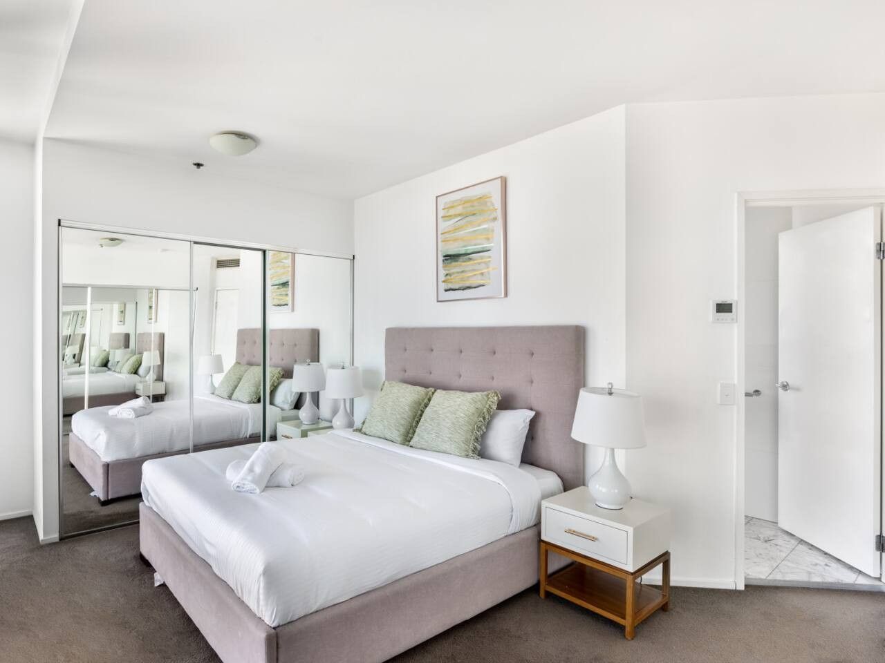 A modern hotel room with a large bed, two bedside tables, a mirror, and a door leading to the bathroom.