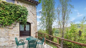 A stone building with a balcony, a table with chairs, and a wooden railing overlooking a forested area.