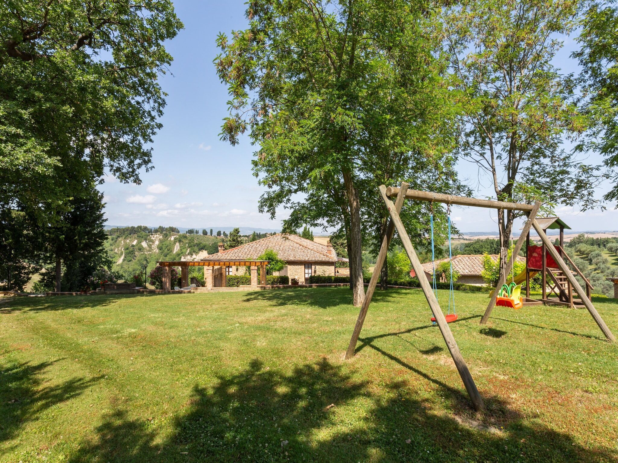 A grassy outdoor area with a wooden swing set, a covered seating area, and a view of a hilly landscape in the background.