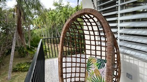 A wicker swing chair with a tropical leaf pattern cushion, attached to a balcony with a black railing, overlooking a garden with palm trees and a white building in the background. - TURQUESA del MAR, private tropical oasis! (Rincon)