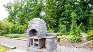 Ein aus Stein gemauerter Außenofen in einem angelegten Garten mit Bäumen und Sträuchern im Hintergrund.