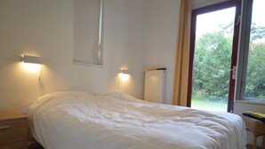 A bedroom with a single bed, a nightstand, a wall-mounted light, and a window with a view of greenery.
