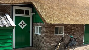 Edifici de maó amb una porta verda i finestres, una bicicleta aparcada al davant i un teulat de palla.