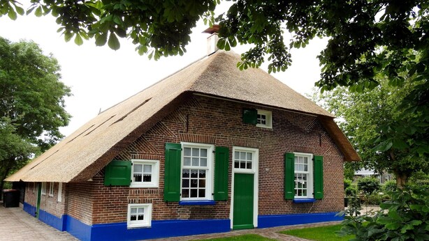 A traditional brick house with a thatched roof, green shutters, and a blue-painted base.