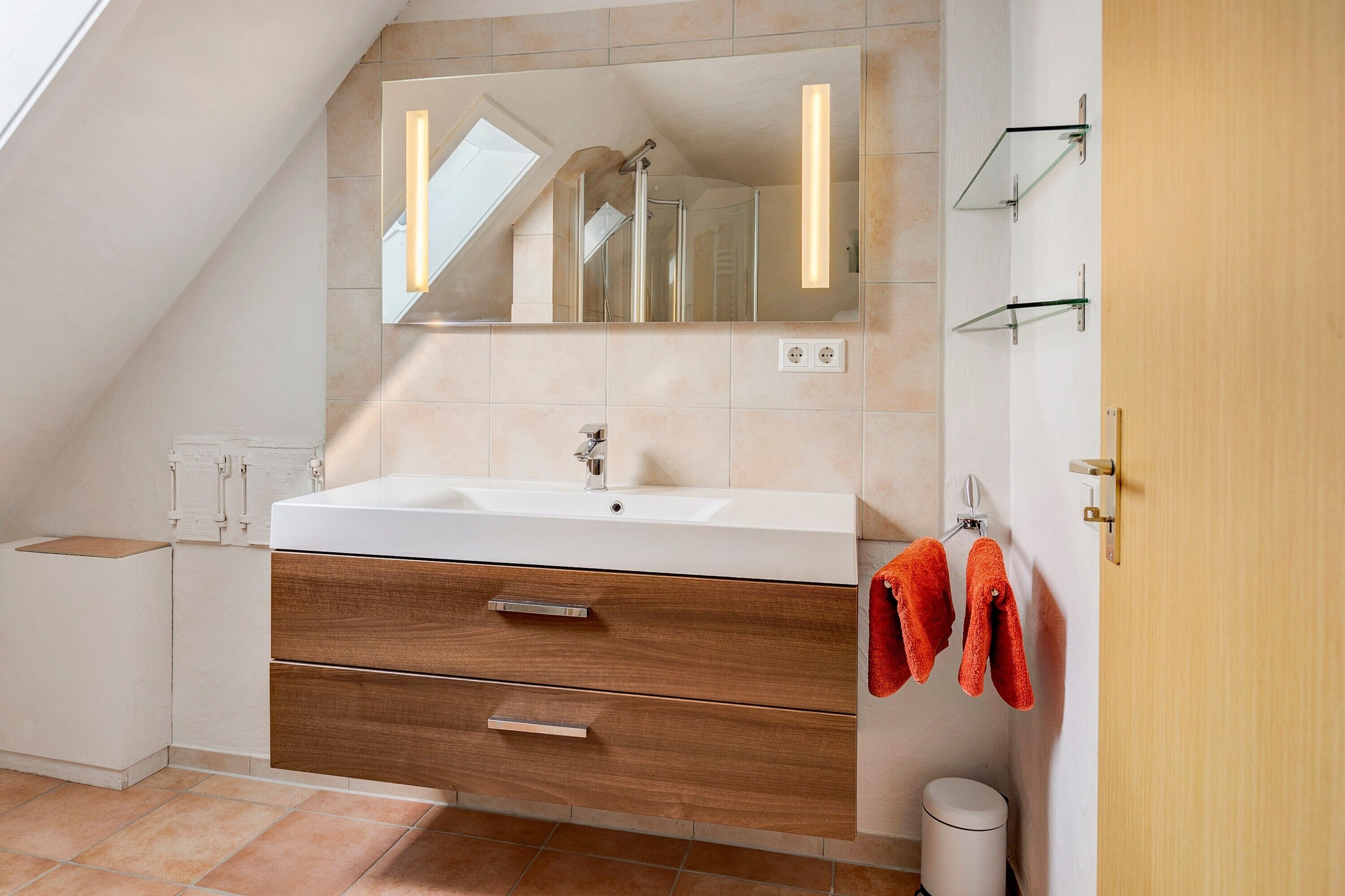 Een moderne badkamer met dakraam, grote spiegel, witte wastafel, houten lades en een handdoekrek met rode handdoeken.