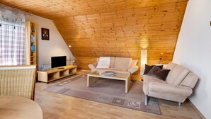 A cozy loft-style room with a wooden ceiling, a sofa, a coffee table, a TV on a wooden stand, and a bookshelf. - Apartment 'Haus Am Südhang' with Shared Garden, Balcony and Wi-Fi (Lenzkirch)
