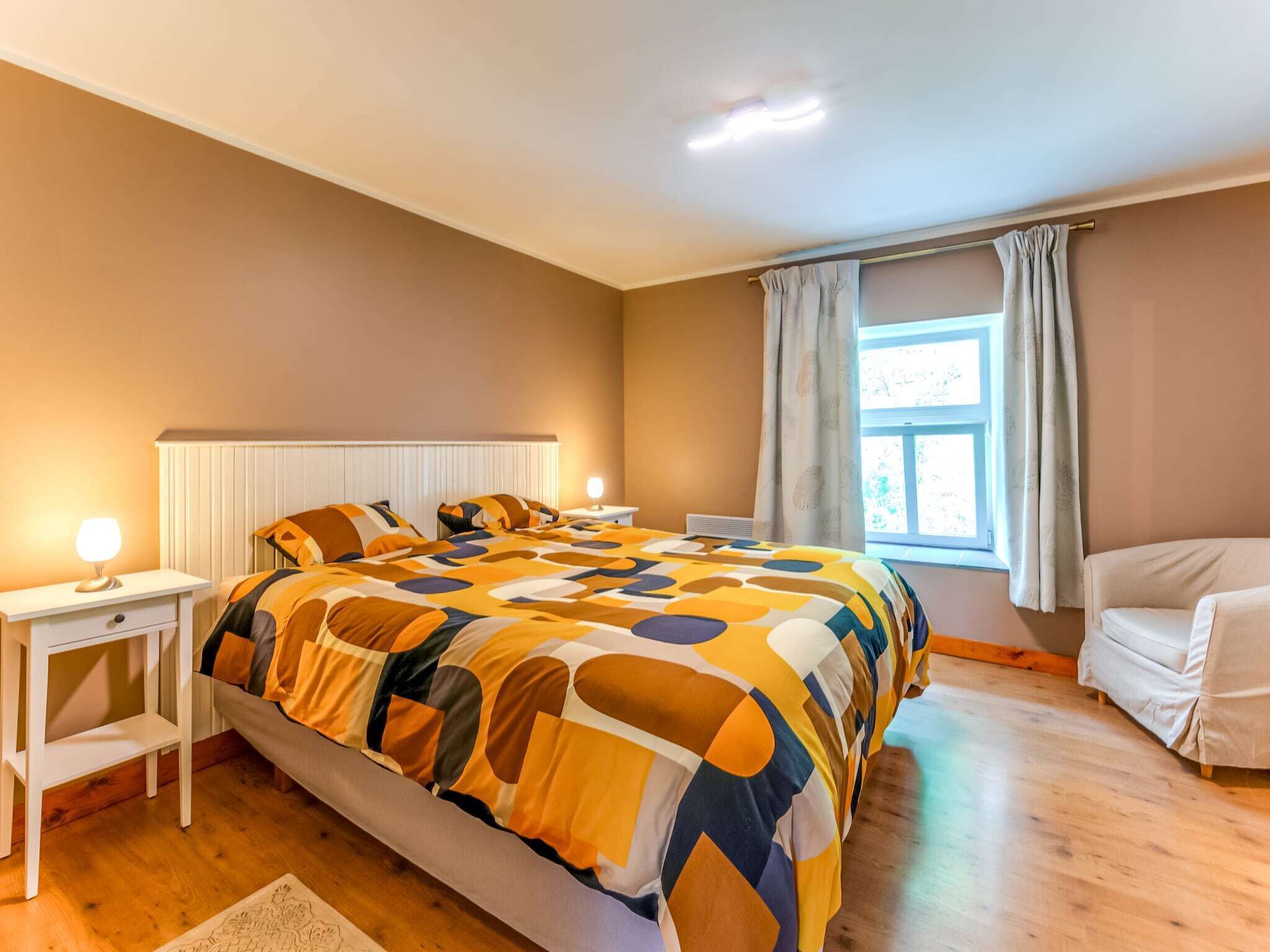 A bedroom with a large bed, a bedside table with a lamp, a window with curtains, and a chair.