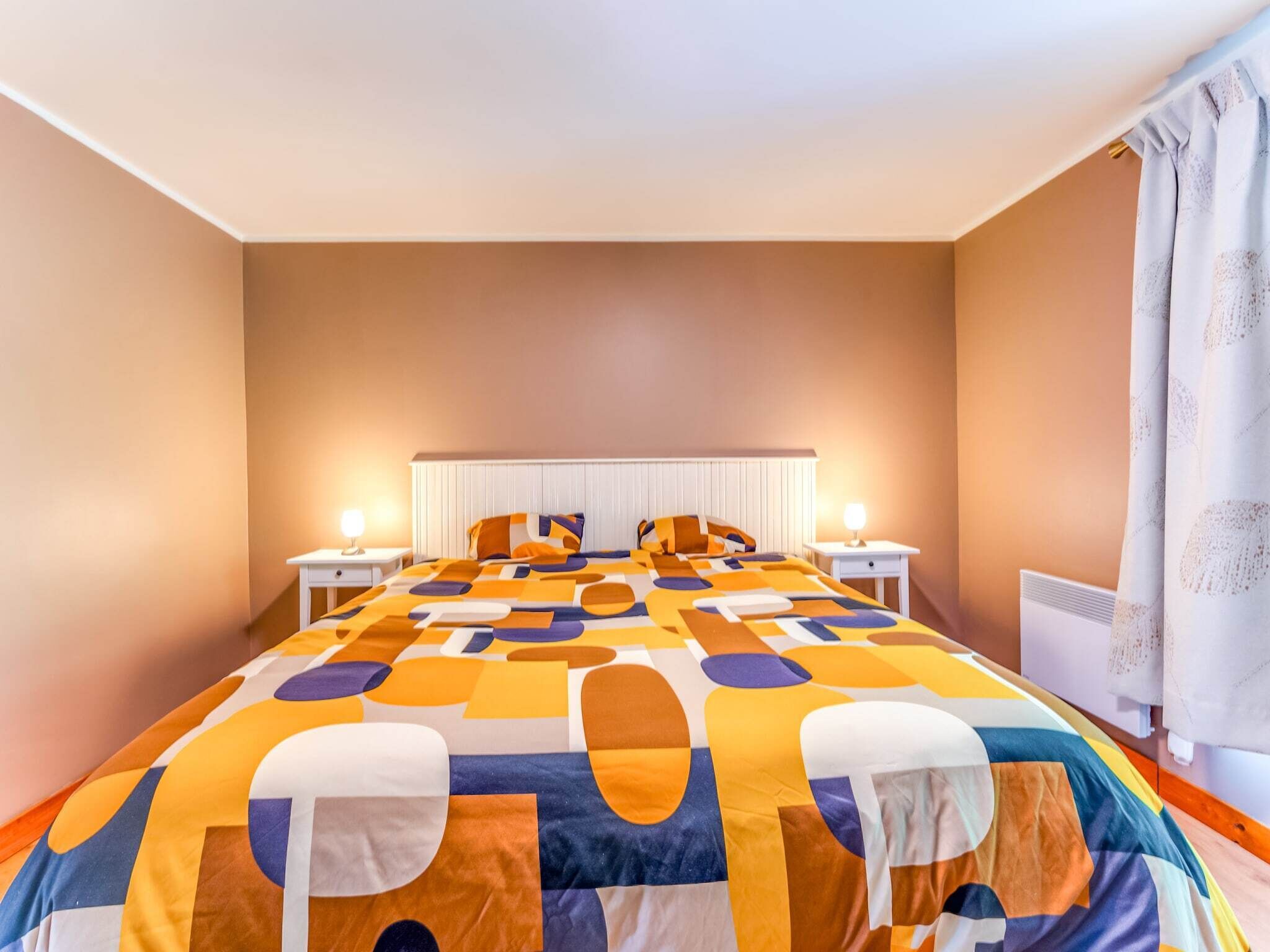 Habitación de hotel con una cama grande, dos mesitas de noche, un radiador y una ventana con cortinas.