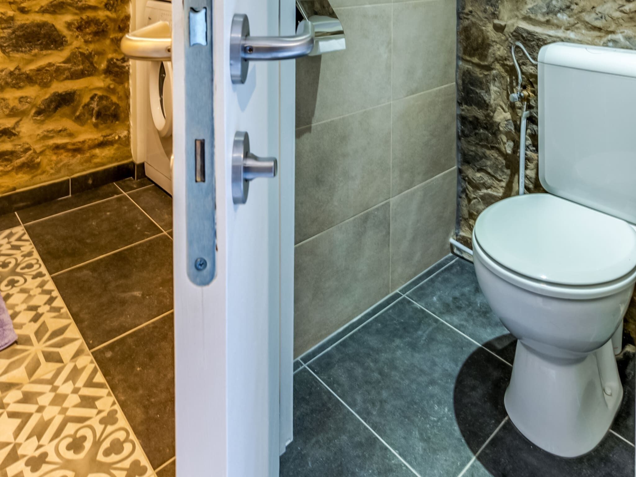 A bathroom with a white toilet, a shower area with a stone wall, and a patterned floor.