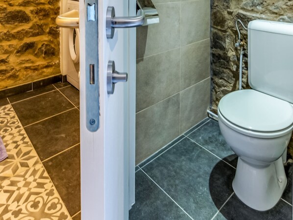 Un baño con inodoro blanco, zona de ducha con pared de piedra y suelo estampado.