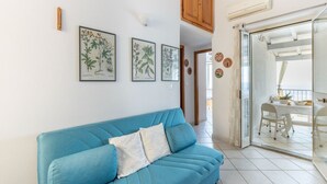 Un salón con un sofá azul, impresiones botánicas enmarcadas en la pared y vistas a un balcón con mesa y sillas.