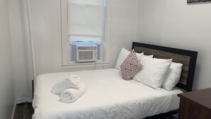 A neatly made bed with white linens and a rolled towel, a window with an air conditioning unit, and a wooden headboard.