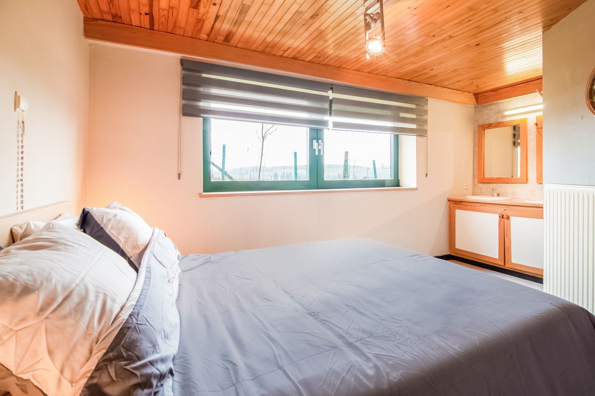 Une chambre à coucher avec un lit, une fenêtre avec des stores, un plafond en bois, une table de chevet et un miroir.