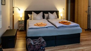 A neatly made double bed with striped bedding, two bedside lamps, a small bedside table with a lamp, and a dark-colored bench. - Apartment 'Moderne Ferienwohnung In Der Stadt' with Balcony and Wi-Fi (Forst/Lausitz)