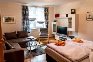 Une chambre d’hôtel moderne avec un canapé, une table basse en verre, un lit recouvert d’une couvre-lit à motifs, une télévision et une bibliothèque.