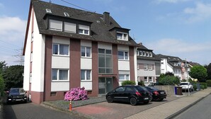 Un edificio residencial de varios pisos con fachada roja y blanca, una entrada principal y un camino de acceso con coches estacionados.