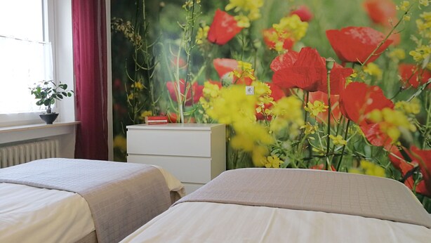 Habitación de hotel con dos camas, una ventana con cortinas rojas, un radiador, un aparador blanco y un mural floral en la pared.