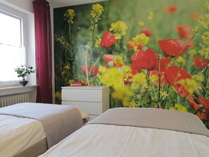 Hotellihuone, jossa on kaksi sänkyä, ikkuna punaisilla verhoilla, patteri, valkoinen lipasto ja seinämaalaus, jossa on kuvattuna kukkia.
