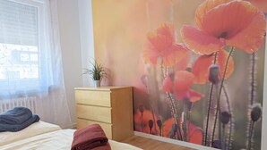 A bedroom with a bed, a wooden dresser, a window with blinds, and a wall with a floral mural. - Holiday Apartment 'Haus Abendsonne Upper Floor Small' with Shared Terrace, Balcony and Wi-Fi (Koblenz)
